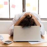 Frustrated person with head in hands at a desk with laptop and papers, symbolising stress and overwhelm at work