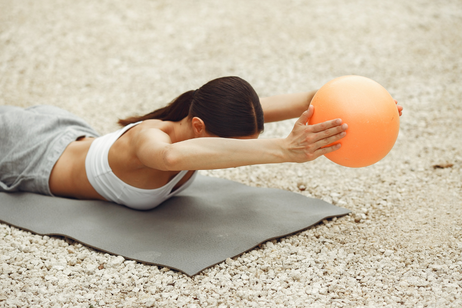 Woman exercising with an orange ball on a mat outdoors on a gravel surface, wearing sportswear for a workout routine