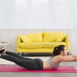 A woman performs a back extension on a pink mat, holding weights in a living room