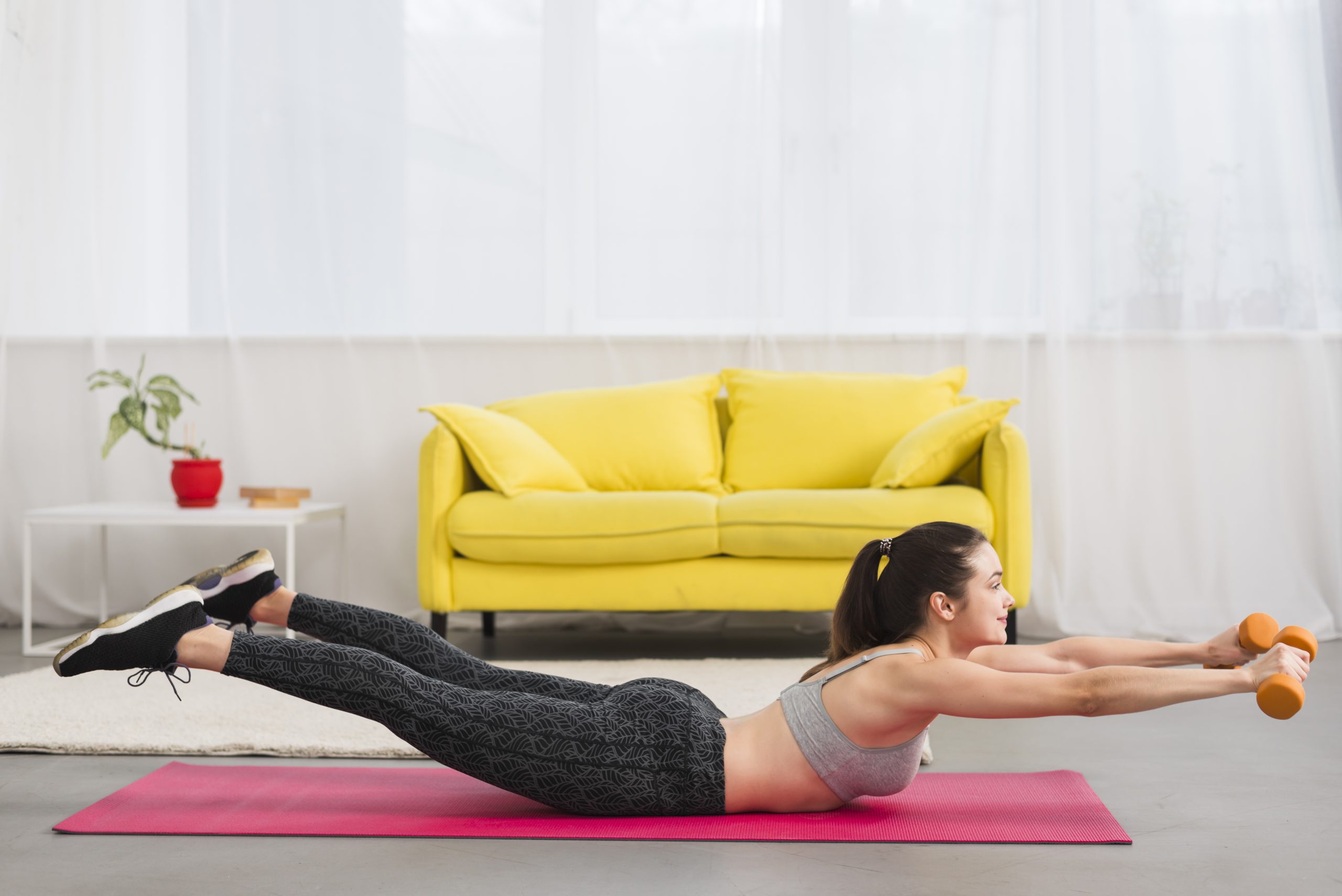 A woman performs a back extension on a pink mat, holding weights in a living room